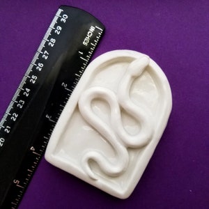 May include: White ceramic mold shaped like an archway with a snake design. The mold is approximately 4 inches wide and 5 inches tall.