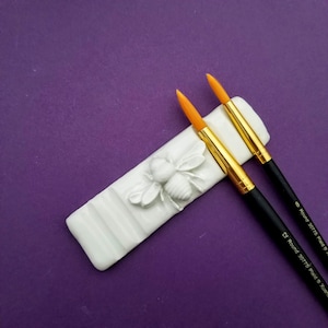 Op de afbeelding: Twee oranje-gekleurde verfkwasten met zwarte handvatten en gouden ferrules liggen in een witte keramische kwastenhouder. De houder heeft een reliëf van een bij en een reeks groeven om de kwasten vast te houden. De kwasten zijn geëtiketteerd met "#0 Round 30775 Plaid" en "#2 Round 30779 Plaid".