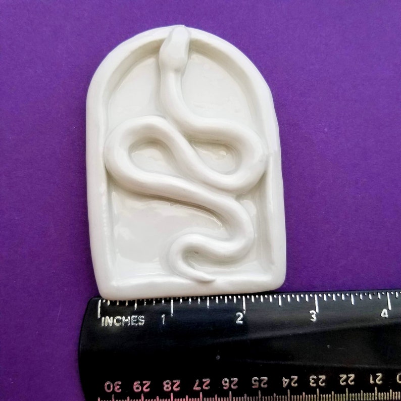May include: White ceramic mold of a snake in a rectangular shape. The mold is approximately 2 inches wide and 3 inches tall.