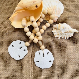 Könnte beinhalten: Eine dekorative Girlande mit hellen Holzperlen und zwei weißen Sanddollar-Anhängern. Die Girlande ist neben einer großen, glatten, blassgelben Muschel, einer weißen fächerförmigen Muschel und einer kleineren, strukturierten weißen Muschel platziert.
