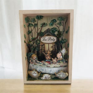 May include: A rectangular wooden shadow box featuring a three-dimensional diorama of a tea party scene. The interior displays a forest backdrop with a sign reading Tea Party, a table with Alice, a rabbit, and a hatter, plus two ceramic teapots.