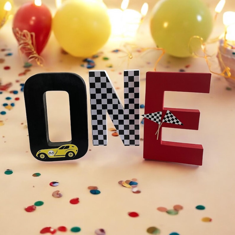 May include: Wooden block letters spelling "ONE" in black, white and red. The "O" has a yellow racing car on it, the "N" has a black and white chequered pattern, and the "E" has two black and white chequered flags.