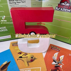 May include: A red and white number 5, with a Poké Ball design, sits in front of Pokémon Trading Card Game materials. The image includes tutorial guides featuring Pikachu and Cinderace, and a game board.