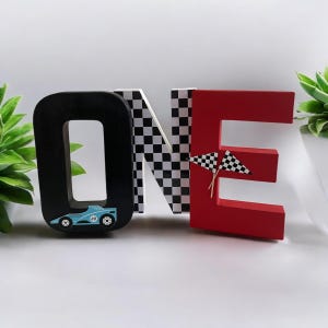 May include: Wooden letters spelling "ONE" in black, white, and red. The "O" has a blue racing car on it, the "N" has a black and white chequered pattern, and the "E" has two red and white chequered flags.