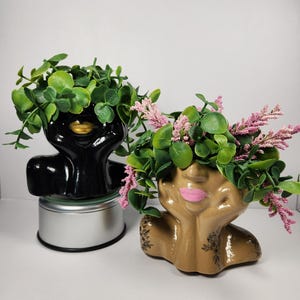 May include: Two ceramic face planters, one black with gold lips, the other tan with pink lips. Both have green foliage and pink accents. The planters are in a resting pose, with hands supporting the face.