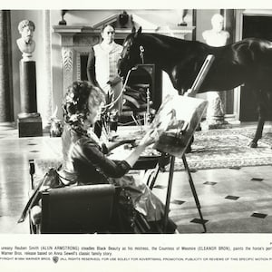May include: A black and white photo of a woman painting a portrait of a black horse. The woman is sitting in a chair and the horse is standing in front of her. The photo is from the movie "Black Beauty".