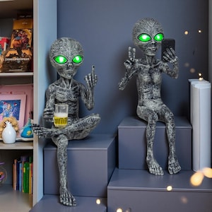 May include: Two gray alien statues with glowing green eyes. One alien is holding a beer and giving the middle finger, while the other is taking a selfie with a smartphone. Both are seated on gray platforms.