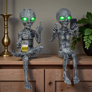 Könnte beinhalten: Zwei graue Alien-Figuren mit leuchtend grünen Augen. Einer hält ein Bier und zeigt den Mittelfinger, der andere macht ein Selfie mit einem Telefon. Die Statuen sitzen auf einer Holzoberfläche.