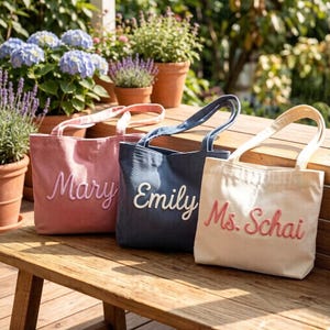 May include: Three personalized tote bags in pink, navy, and cream, each with a name embroidered in a contrasting color. The bags sit on a wooden bench, with potted plants in the background. The names are "Mary", "Emily", and "Ms. Schai".