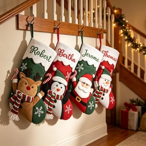 Könnte beinhalten: Vier Weihnachtsstrümpfe hängen in einer Reihe, jeder mit einer weißen Manschette und den eingestickten Namen Robert, Bertha, Jerome und Thomas. Die Strümpfe zeigen festliche Figuren wie ein Rentier, einen Pinguin, einen Weihnachtsmann und einen Schneemann.