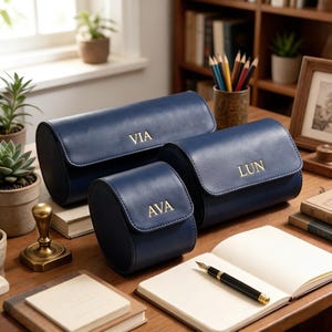 May include: Three navy blue leather watch cases with gold lettering spelling "VIA", "LUN", and "AVA". The cases are cylindrical with a flap closure. They are arranged on a wooden desk with a notebook and pen.