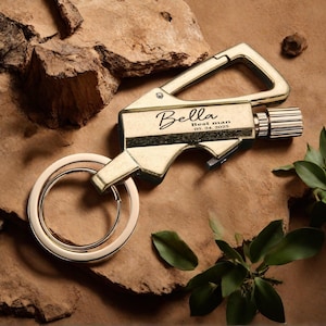 May include: A gold-colored keychain with a carabiner clip, a bottle opener, and a key ring. The keychain has the name "Bella" and the text "Best man 05. 24. 2025" engraved on it. The background is a textured brown surface with green leaves.