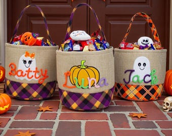 Puede incluir: Tres cestas cilíndricas de tela con asas, con cuerpos de arpillera color canela y bases a cuadros en morado, naranja y negro. Cada cesta muestra una aplicación de fieltro, un fantasma o una calabaza, y nombres bordados: Batty, Luna y Jack.
