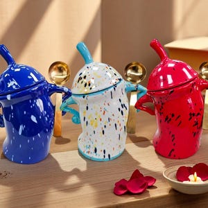 Puede incluir: Tres azucareros de cerámica en azul, blanco y rojo, cada uno con tapa a juego y cuchara dorada. El cuenco blanco tiene salpicaduras de pintura de colores. Los cuencos están sobre una superficie de madera con un cuenco pequeño de pétalos de rosa y una vela.