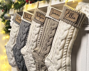 以下が含まれることがあります： 白、グレー、ベージュの色合いのケーブルニットのクリスマスストッキングが5つ、白いマントルピースに吊るされています。「Charley」や「Ellie」などの名前が書かれた木製のネームタグが付いています。マントルピースはクリスマスのガーランドで飾られています。