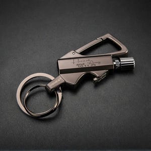 May include: A gunmetal-colored keychain lighter with a carabiner clip and a key ring. The lighter has the word "HONEST" printed on it and the inscription "Hannah and 10.11.25". The lighter has a textured, ridged wheel for ignition.