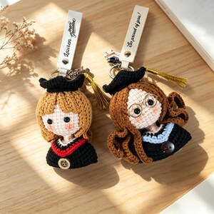 May include: Two handmade crochet graduation keychains. One keychain features a blonde-haired doll wearing a black graduation gown with a red and black collar. The other keychain features a brown-haired doll wearing a black graduation gown with a blue and black collar. Both keychains have a tassel.