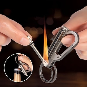 May include: A metallic gray keychain lighter with a carabiner clip and key rings. The lighter is lit, showing a flame. The image also shows the lighter opening a bottle cap. The lighter is held by a person's hands.