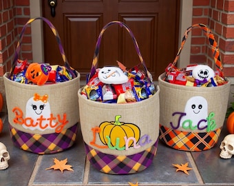Puede incluir: Tres cestas cilíndricas de Halloween hechas de tela color canela con bases de cuadros morados y naranjas. Cada cesta tiene un asa, un aplique de fieltro de fantasma o calabaza y los nombres bordados: Batty, Luna y Jack. Están llenas de dulces.