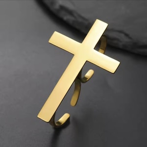 May include: A gold-colored cross-shaped bracelet. The bracelet features a simple, elegant design with a polished finish. The cross is the focal point, with a smooth, reflective surface. The bracelet is open-ended, allowing for easy wear.