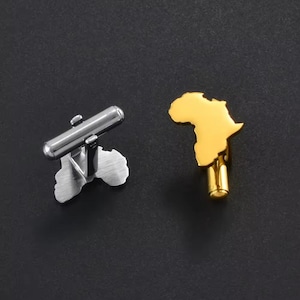 May include: A pair of cufflinks, one silver and one gold, each shaped like the continent of Africa. The silver cufflink has a polished, cylindrical bar. The gold cufflink has a shiny, reflective surface. The background is a dark, solid color.