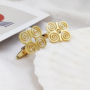 May include: A pair of gold-colored cufflinks with an openwork design featuring a four-leaf clover shape with spiral details. The cufflinks are displayed on a white surface, with a blurred image in the background.