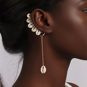 May include: A gold-coloured ear cuff and earring set with white seashells. The ear cuff curves along the ear, while the earring hangs down with a single shell pendant. The jewellery is worn on a person's ear.