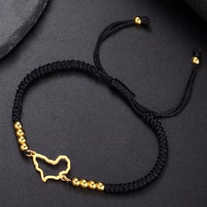 May include: Black braided bracelet with gold-colored beads and a gold-colored charm in the shape of a map. The bracelet has an adjustable sliding knot closure for a comfortable fit.