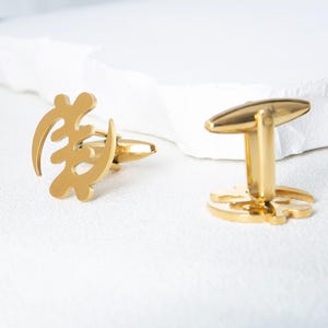 May include: A pair of gold-colored cufflinks. One cufflink features a stylized symbol, while the other has a simple, rounded design. The cufflinks are displayed on a white surface.