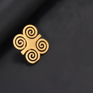 May include: A gold-colored, four-lobed brooch with a central square and four spiral designs. The brooch is set against a dark background, highlighting its bright, reflective surface. The design is symmetrical and geometric.