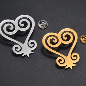 May include: Two heart-shaped pins, one silver and one gold, with a decorative swirl design. Each pin has a small, round backing. The pins are displayed on a dark surface.