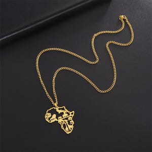 May include: Gold-colored necklace featuring a pendant in the shape of the African continent. The pendant has intricate cut-out details. The chain is a curb style, and the necklace is displayed on a dark surface.