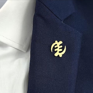 May include: A gold-coloured lapel pin with a unique symbol is attached to a navy blue blazer. The pin is positioned near the lapel, next to a white collared shirt. The symbol is a stylized, abstract design.
