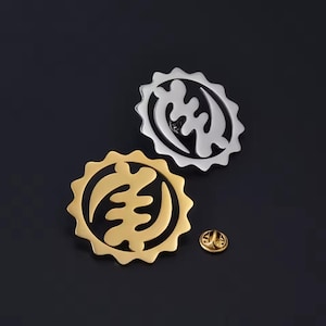 May include: Two metal pins with a sunburst design. One pin is gold-coloured, the other is silver. A small gold-coloured pin is also visible. The pins feature a symbol, likely of cultural significance. The background is a dark, solid colour.