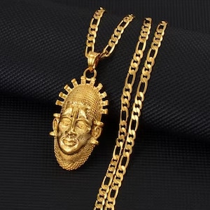 May include: Gold-colored pendant necklace featuring a detailed mask design. The pendant is suspended from a matching gold chain. The necklace is displayed against a dark, textured background, highlighting the jewelry's intricate details.