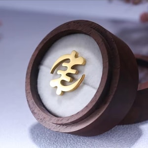 May include: A gold-colored pendant with a unique symbol, displayed inside a round, dark wooden box with a white interior. The pendant is the focal point, set against the box's rich wood grain.