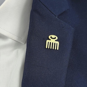 May include: A gold-coloured comb-shaped lapel pin is attached to a navy blue blazer. The pin is on the lapel, near a white collared shirt. The pin has a simple, minimalist design. The blazer is a dark shade of blue.