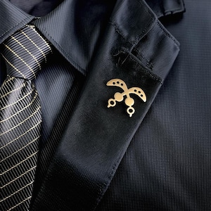 May include: A gold-colored lapel pin in the shape of a stylized bird is pinned to the pocket of a dark blue suit jacket. The jacket is paired with a black tie with a gold grid pattern. The pin has a unique, symmetrical design.