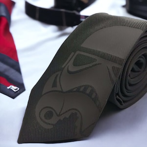 Puede incluir: Una corbata verde oscuro con un gráfico en blanco y negro de un personaje de Star Wars. La corbata está hecha de una tela texturizada y tiene un extremo enrollado.
