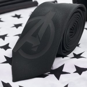 Puede incluir: Corbata negra con un logotipo de los Vengadores en blanco y negro. La corbata está sobre un fondo blanco con estrellas negras.