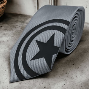 Puede incluir: Una corbata gris con un diseño de estrella y círculo negro. El diseño se asemeja al logotipo de un superhéroe.