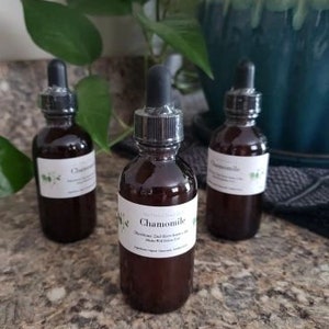 May include: Three dark brown glass bottles with dropper tops filled with a clear liquid. The bottles are labelled "Chamomile" and have a white background with green floral designs.