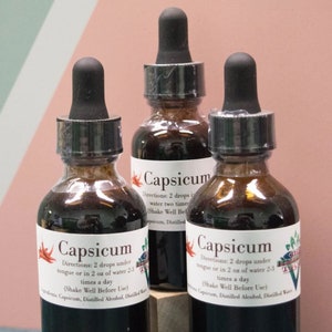 Puede incluir: Tres botellas de vidrio con goteros llenos de un líquido marrón oscuro. Cada botella tiene una etiqueta blanca con la palabra "Capsicum" e instrucciones de uso. Las botellas están sobre una superficie de madera.