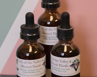 May include: Three amber glass bottles with dropper tops filled with a brown liquid. Each bottle has a white label with the text "Lily of the Valley & Blessed Thistle" and directions for use. The bottles are sitting on a green surface.