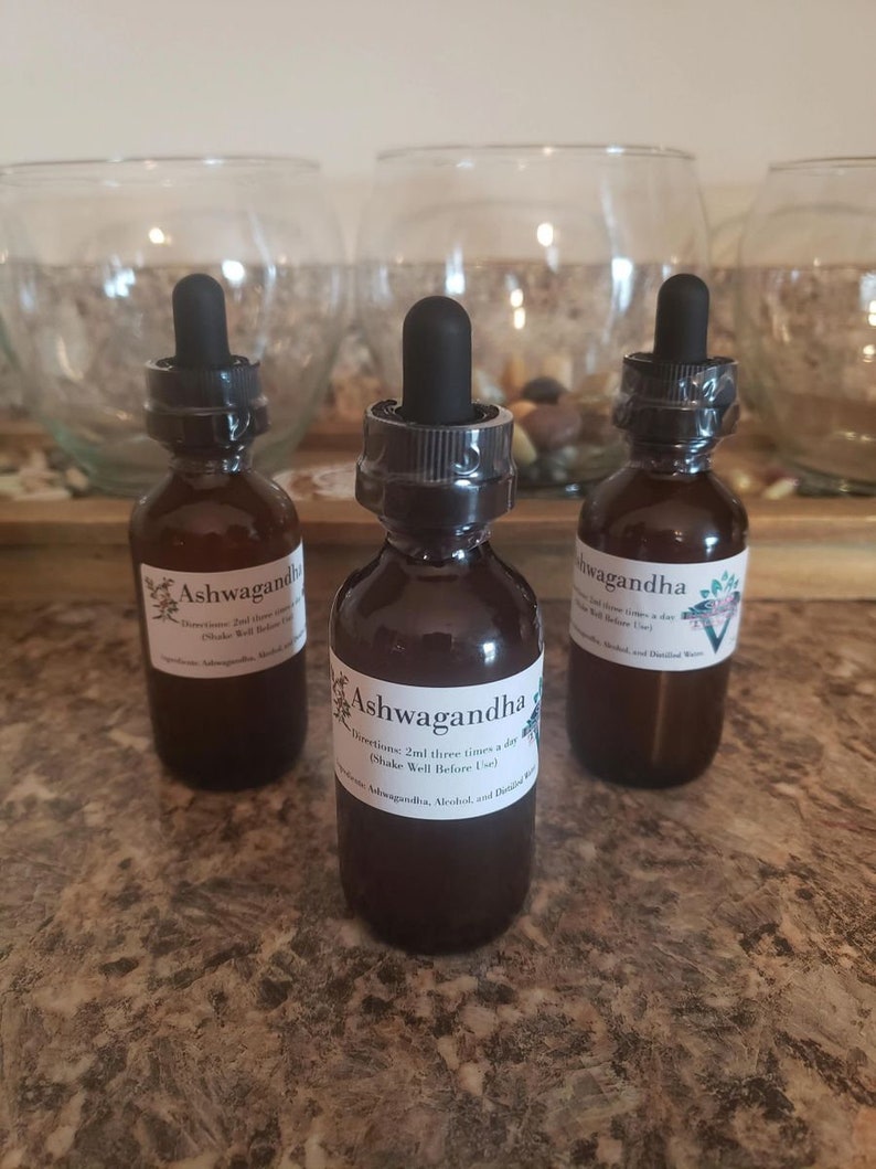 May include: Three brown glass bottles with black droppers, labeled "Ashwagandha." Each bottle has a white label with text. The bottles are arranged on a speckled countertop, with glass bowls in the background.