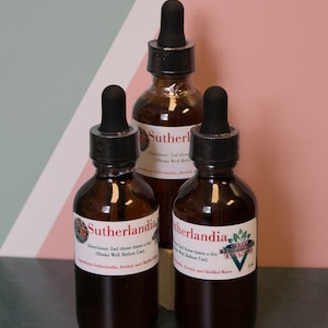 May include: Three brown glass bottles with black droppers. Each bottle has a white label with the word "Sutherlandia" and other text. The bottles are filled with a dark liquid. The background is a pink and white striped wall.