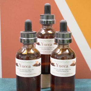 Könnte beinhalten: Drei braune Glasflaschen mit Tropfern, beschriftet mit "The Herbal Touch LLC Yucca" und Anweisungen, 2 ml dreimal täglich einzunehmen. Die Flaschen stehen auf einer Holzoberfläche vor einem Hintergrund aus Orange, Rot und Weiß.