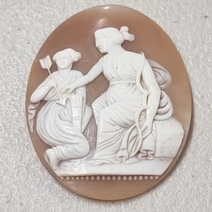 May include: A cameo carved from shell depicting two figures. The figures are a woman holding a bow and arrow and a woman kneeling with a quiver of arrows. The cameo is set in a brown oval frame.