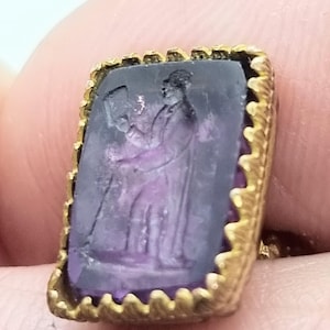 May include: A gold-toned ring with a purple stone featuring a carved image of a person. The stone is set in a decorative gold frame.