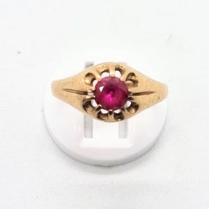 Puede incluir: Un anillo dorado con una gema roja brillante en el centro. El anillo tiene un diseño calado y ornamental alrededor de la gema. El anillo se muestra sobre un soporte blanco.